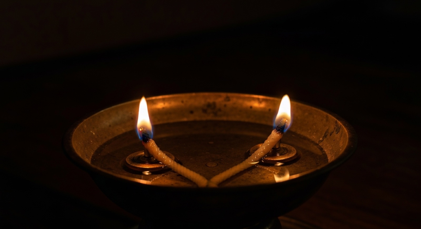 An old brass oil lamp with two wicks set side by side in the same shallow bowl, both alight.
