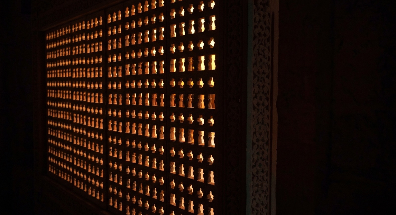 A dense carved stone jali seen at an angle from the dark side. Warm light behind it arrives only as small bright points through the openings; the space beyond is unreadable.